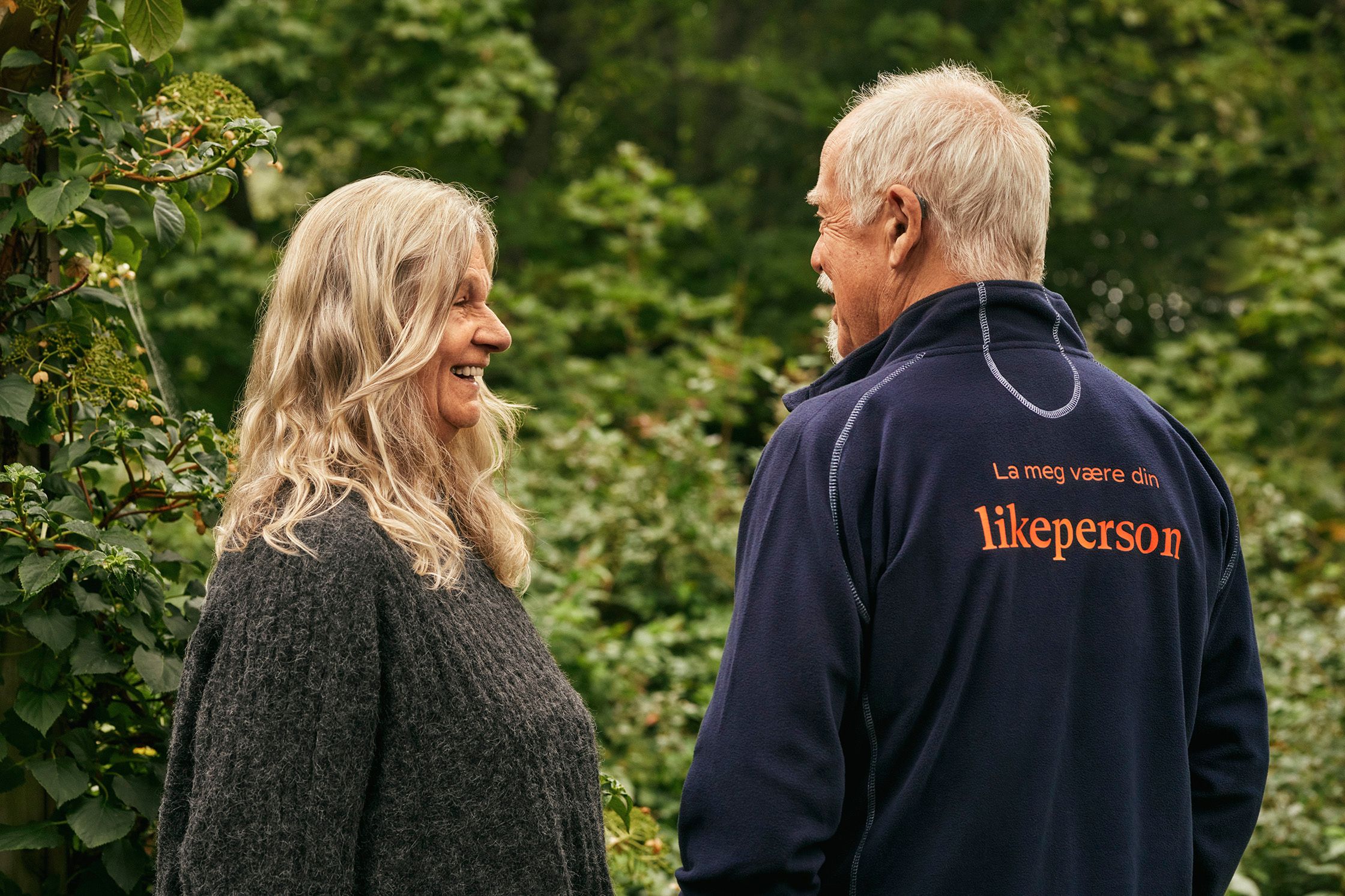 En kvinne og en mann står ved siden av hverandre og smiler. Han har på seg en jakke med teksten La meg være din likeperson.